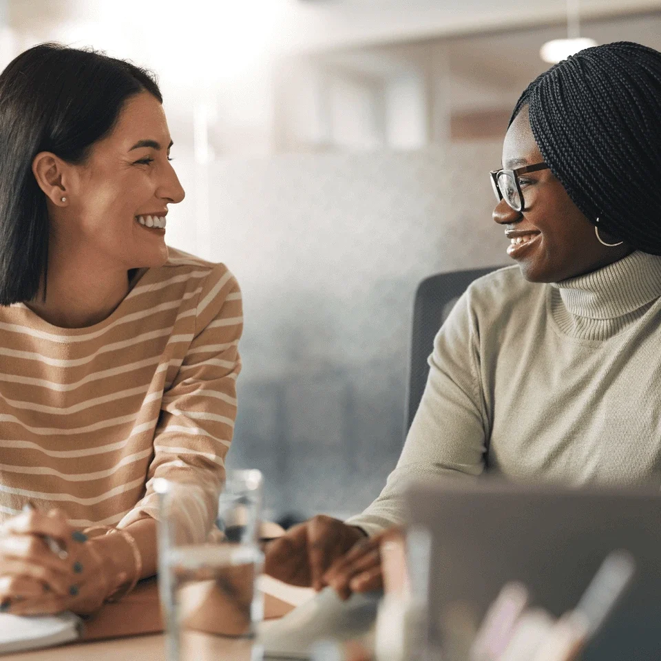 Two women are smiling and engaging in conversation at a desk.