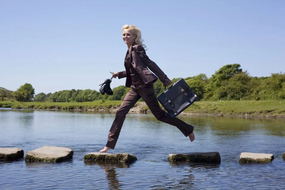 A woman in a suit crosses stepping stones over a river, holding a briefcase and shoes.