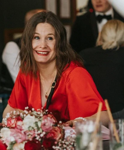 A woman in a red dress smiling, seated at a table, with a wedding bouquet.